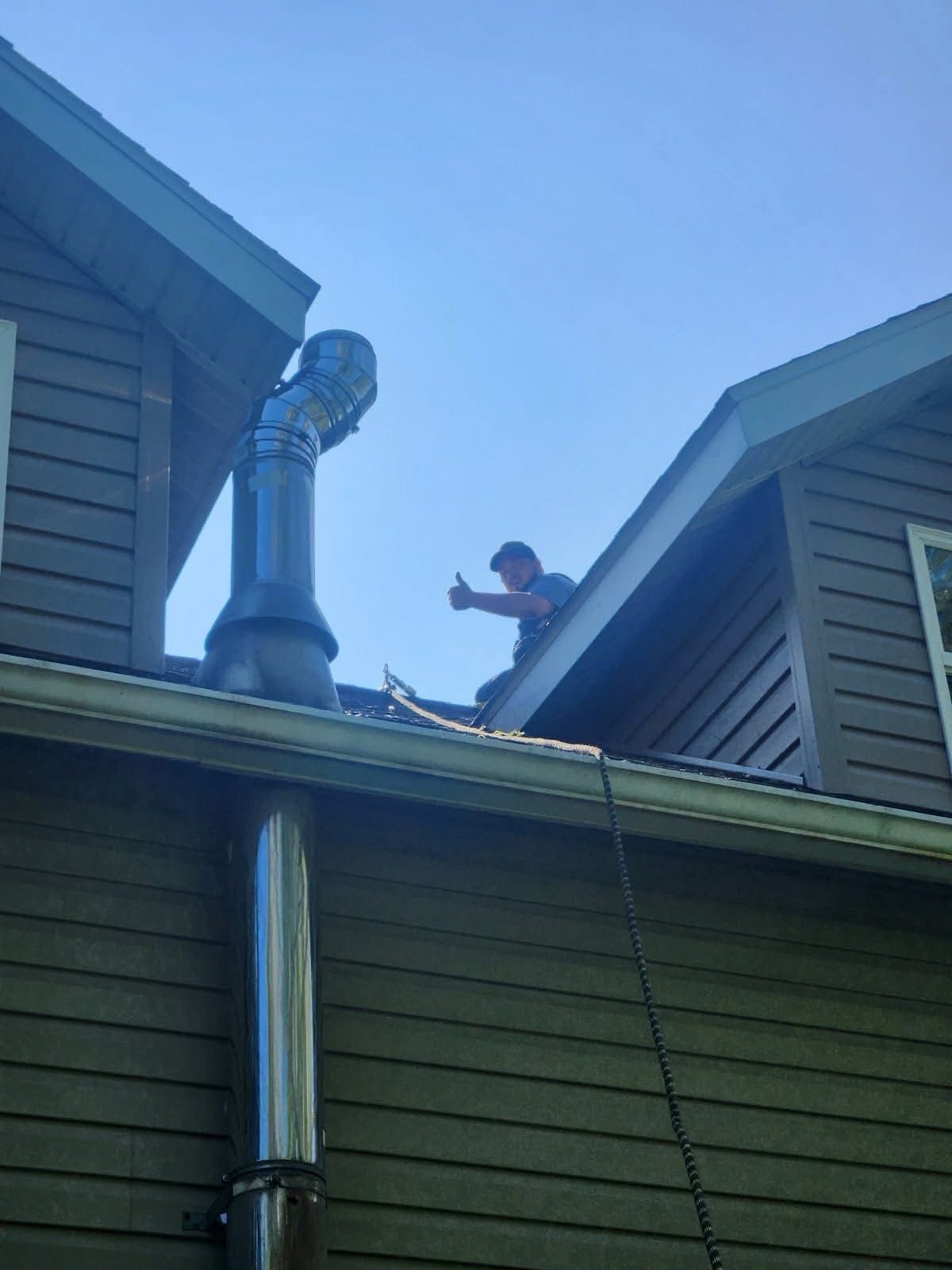 Man on a roof giving a thumbs up near a black chimney against a blue sky.