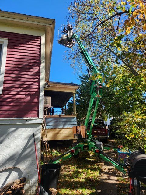 Green lift platform trimming a tree near a red house with a porch on a sunny day.