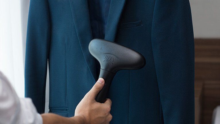 A hand uses a handheld garment steamer on a dark blue suit jacket hanging indoors.