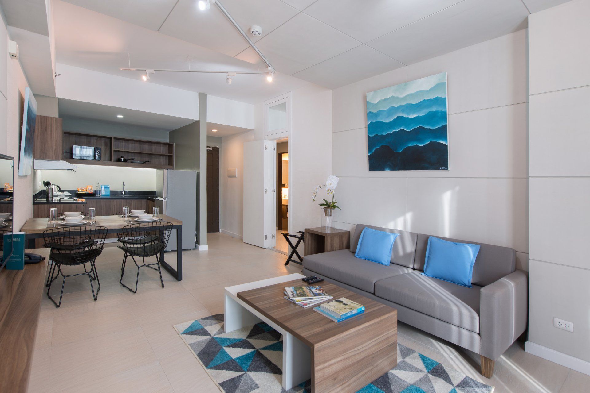Modern open-plan living room with gray sofa, blue accents, coffee table, and kitchen/dining area in the background