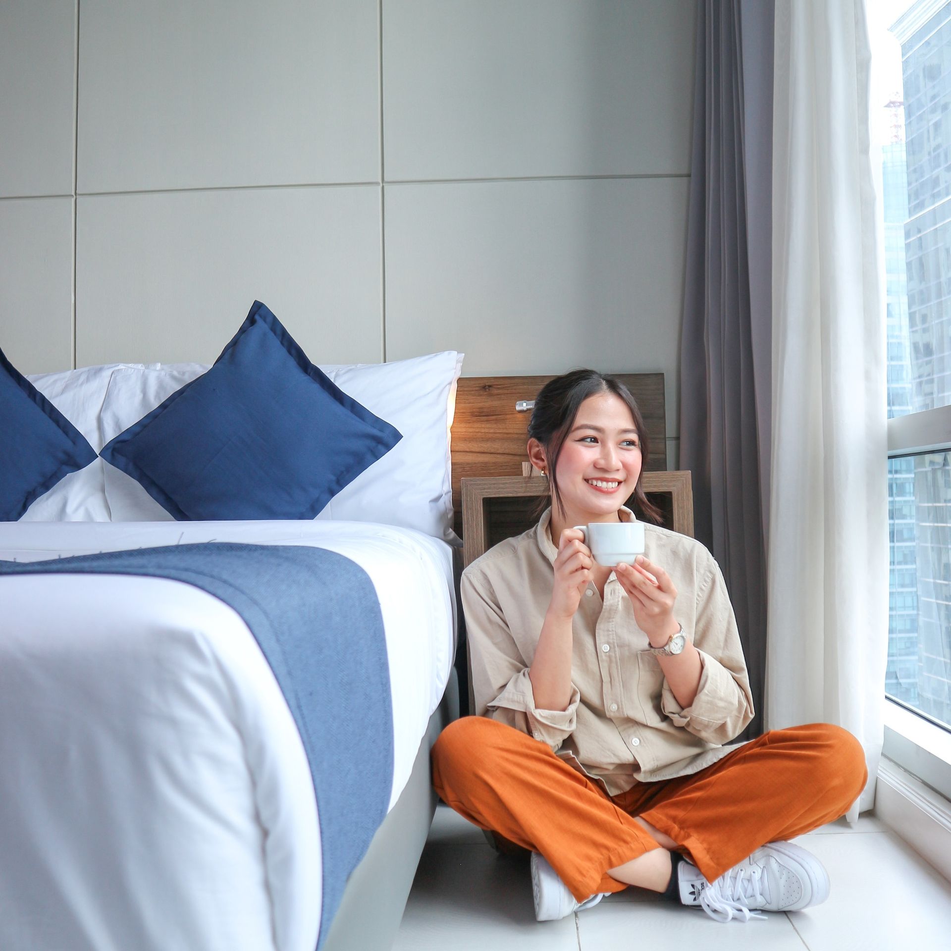 Woman smiles, holding a cup, sitting on floor by a window. Beside her is a bed with blue pillows.