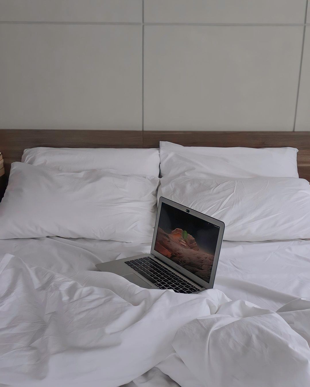Laptop on a bed with white sheets and pillows, against a plain wall.