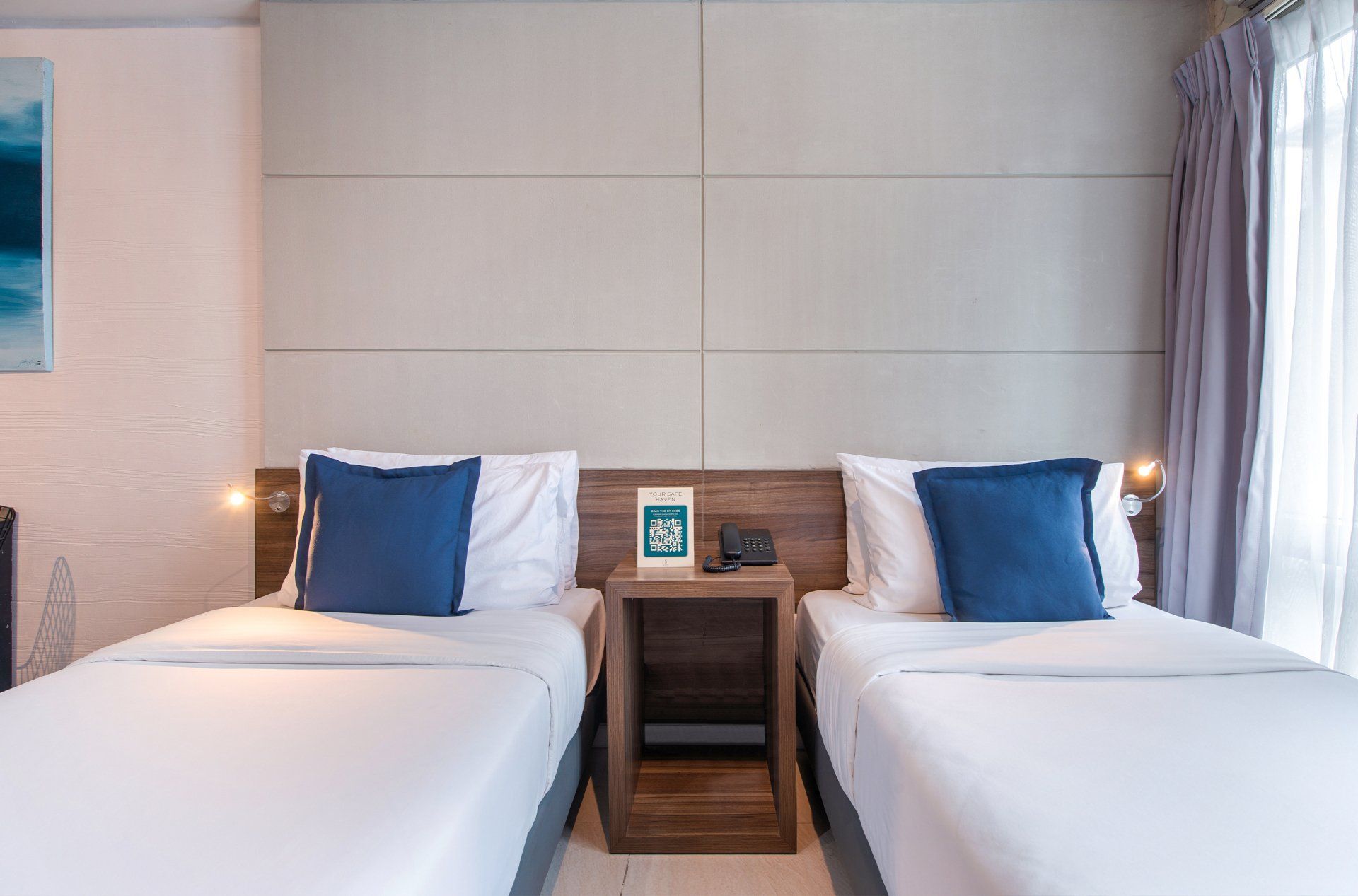 Two twin beds in a hotel room with white bedding and blue pillows, a wooden nightstand.
