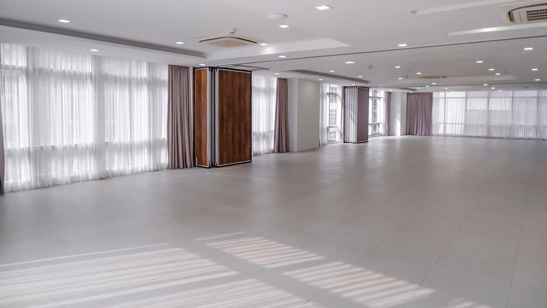Empty, spacious white room with sheer curtains, wooden panels, and recessed lighting. Sunlight casts shadows on floor.