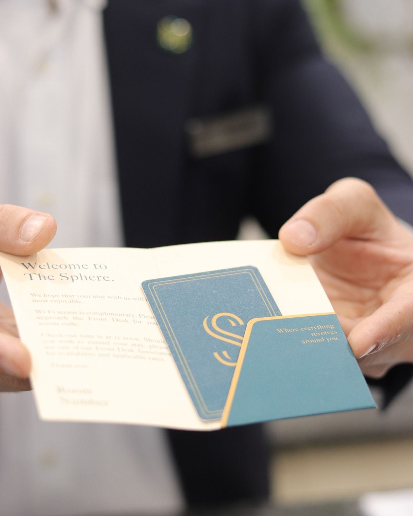 A person in a dark blazer presents a welcome card and a teal key card branded with the letter S for The Sphere.