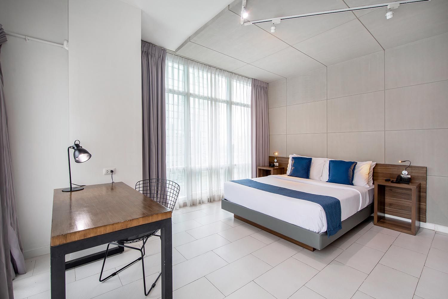 A modern hotel room with a queen bed, a wood-topped desk, white tile floors, and large windows with gray curtains.
