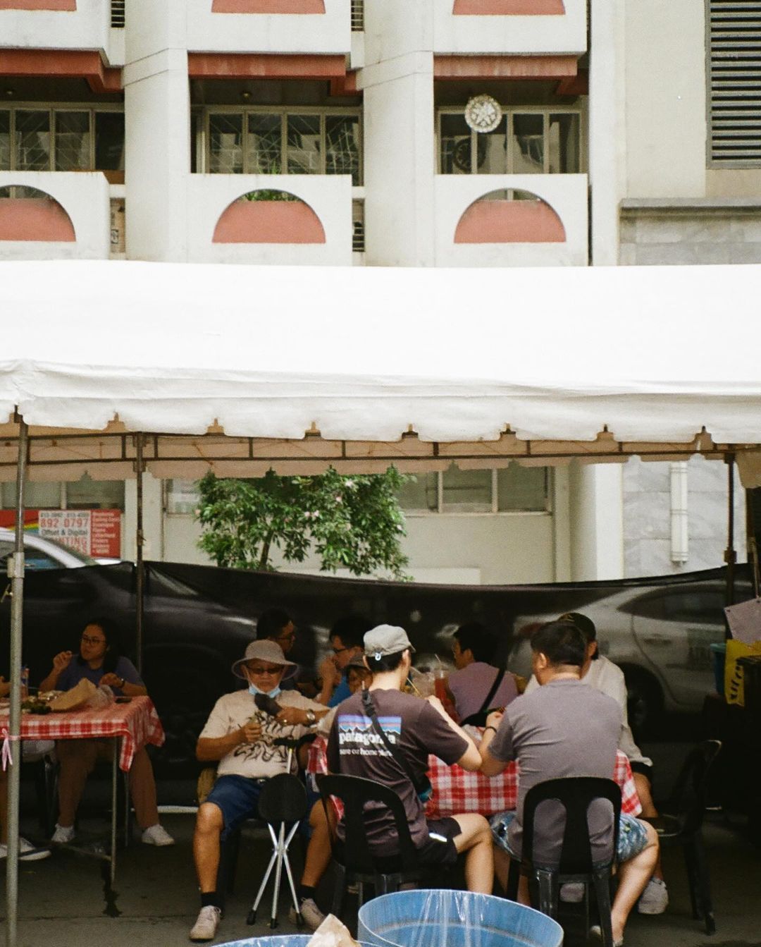 People eating at an outdoor table under a white tent. Building with arched windows in the background.