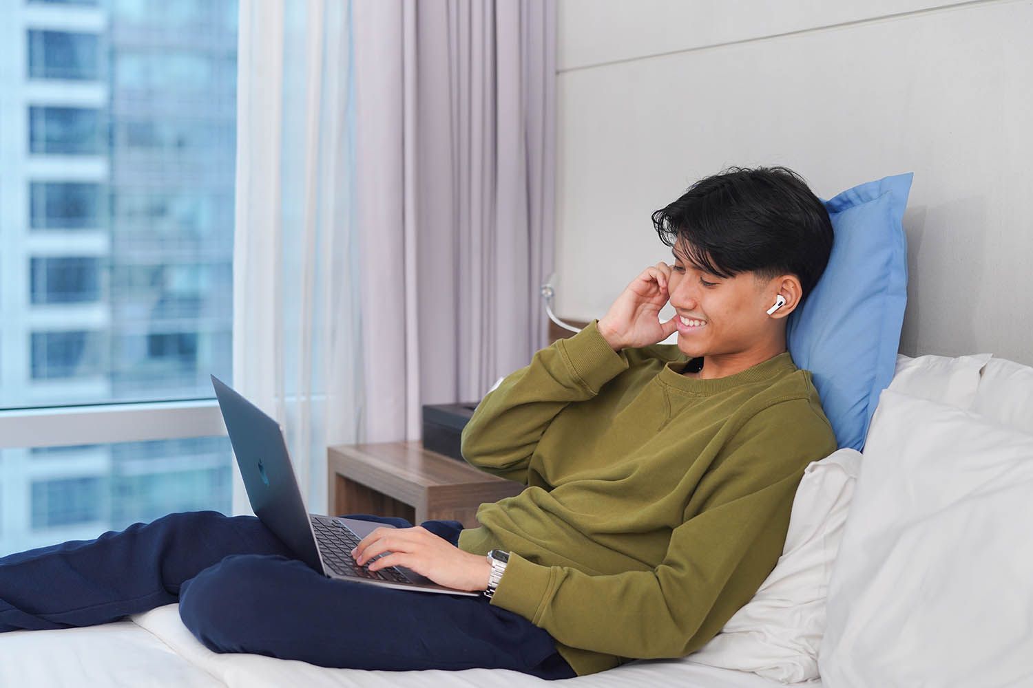Person on bed with laptop and earbuds, smiling. Bright room, cityscape view.