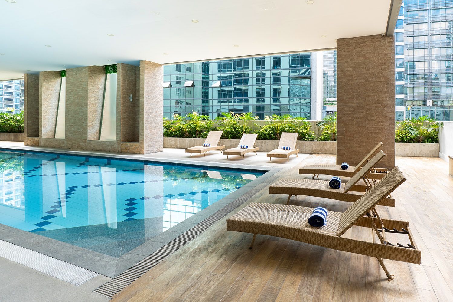 Indoor pool with lounge chairs, city view. Blue water, wooden deck, tan chairs.