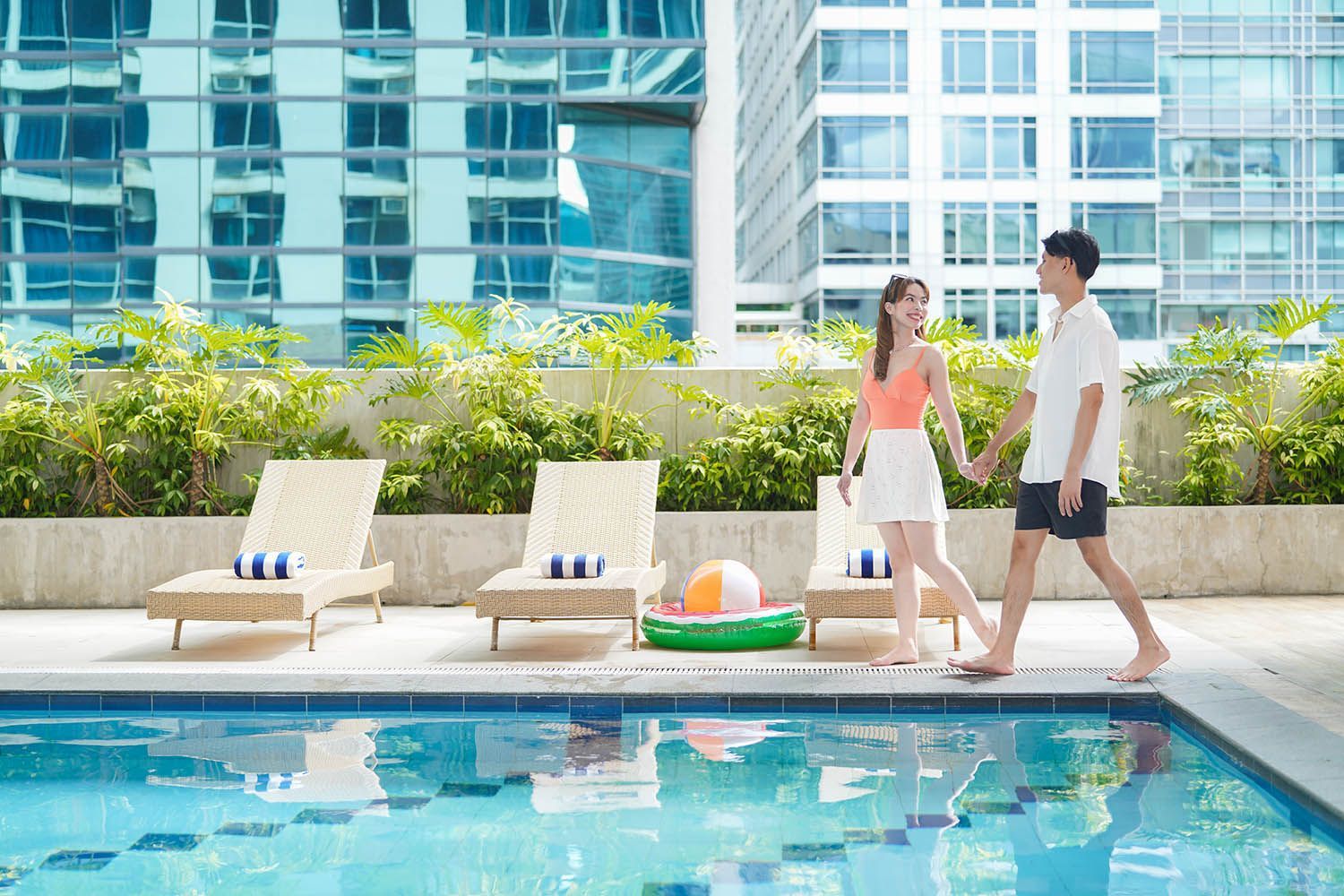 Couple holding hands by a pool, walking toward a building. Suntanning chairs and a floaty are present.