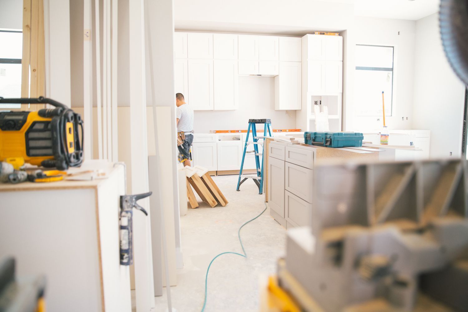 Kitchen under construction, light cabinets, blue ladder, worker installing, power tools, unfinished walls, bright.