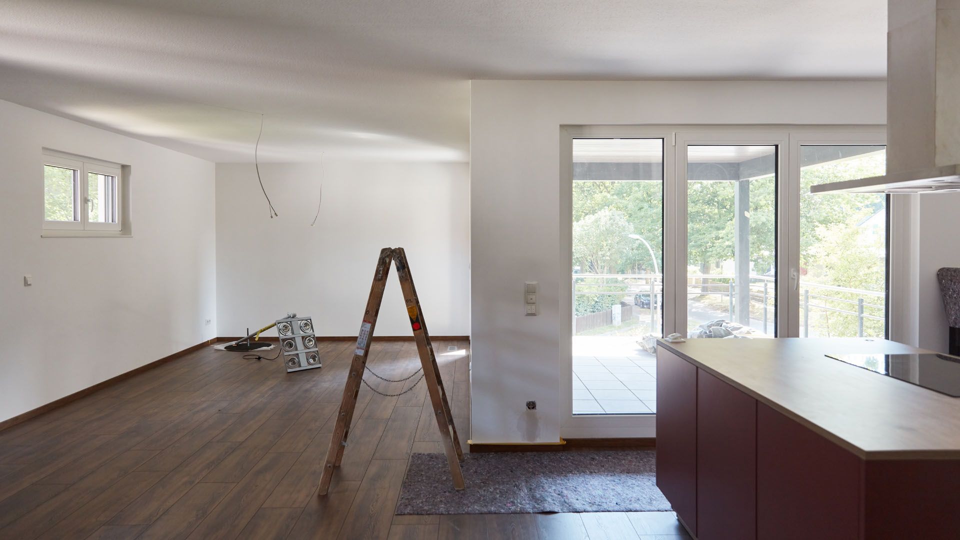 Interior of a room under renovation with hardwood floors, a ladder, and large windows.