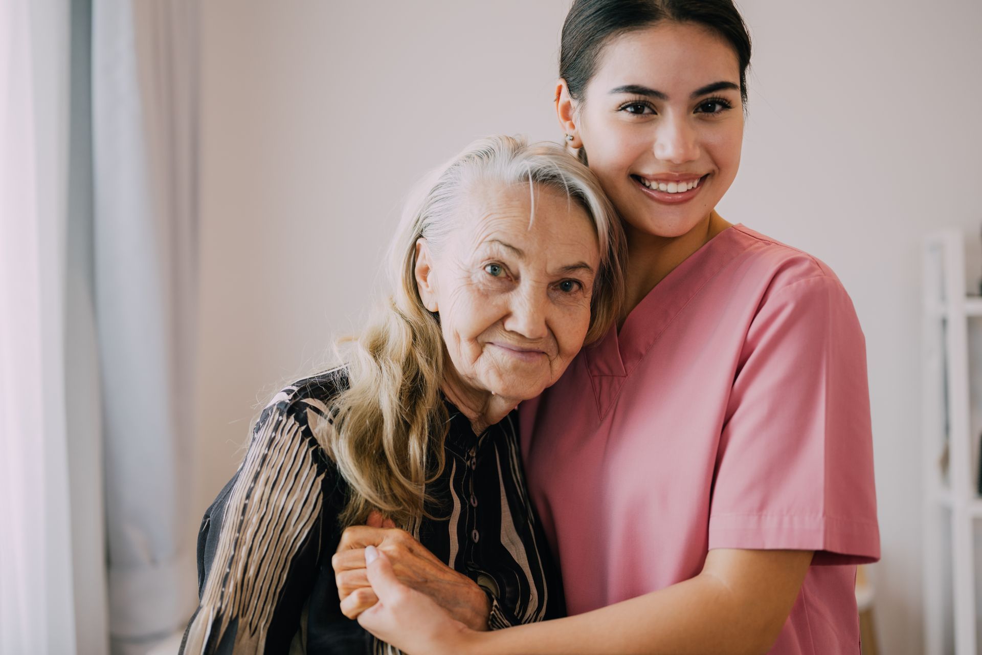 Young woman in pink scrubs hugging an elderly woman, both smiling indoors.