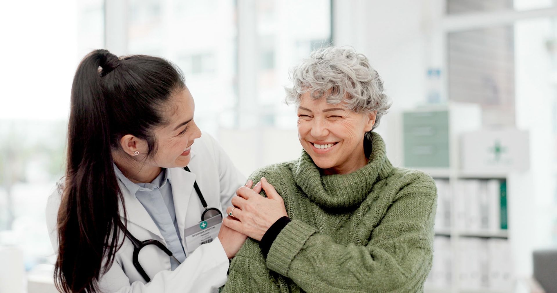 Doctor with a stethoscope, arm around a smiling patient in a green sweater. Bright, indoor setting.