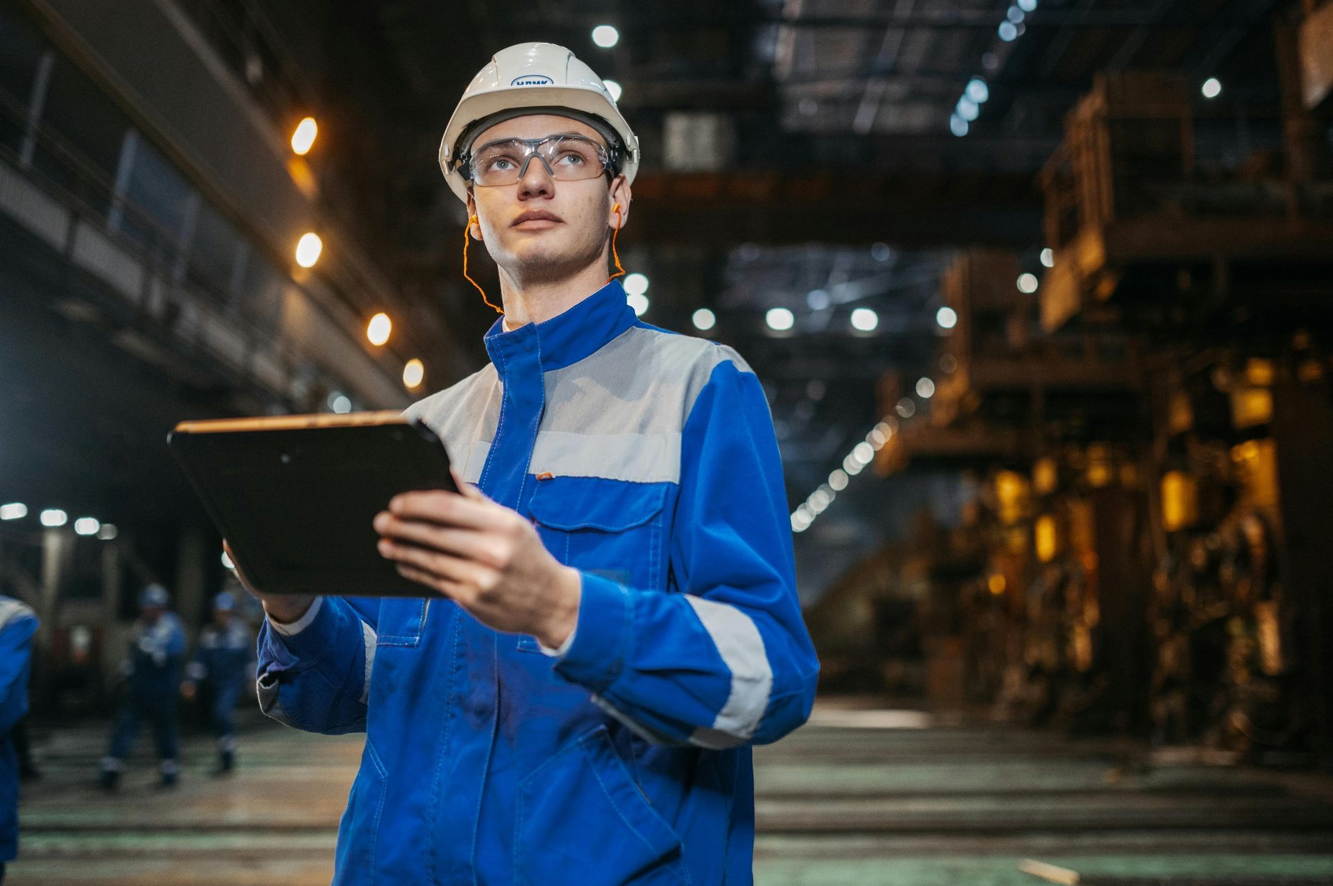 A man in a hard hat is holding a tablet in a factory.