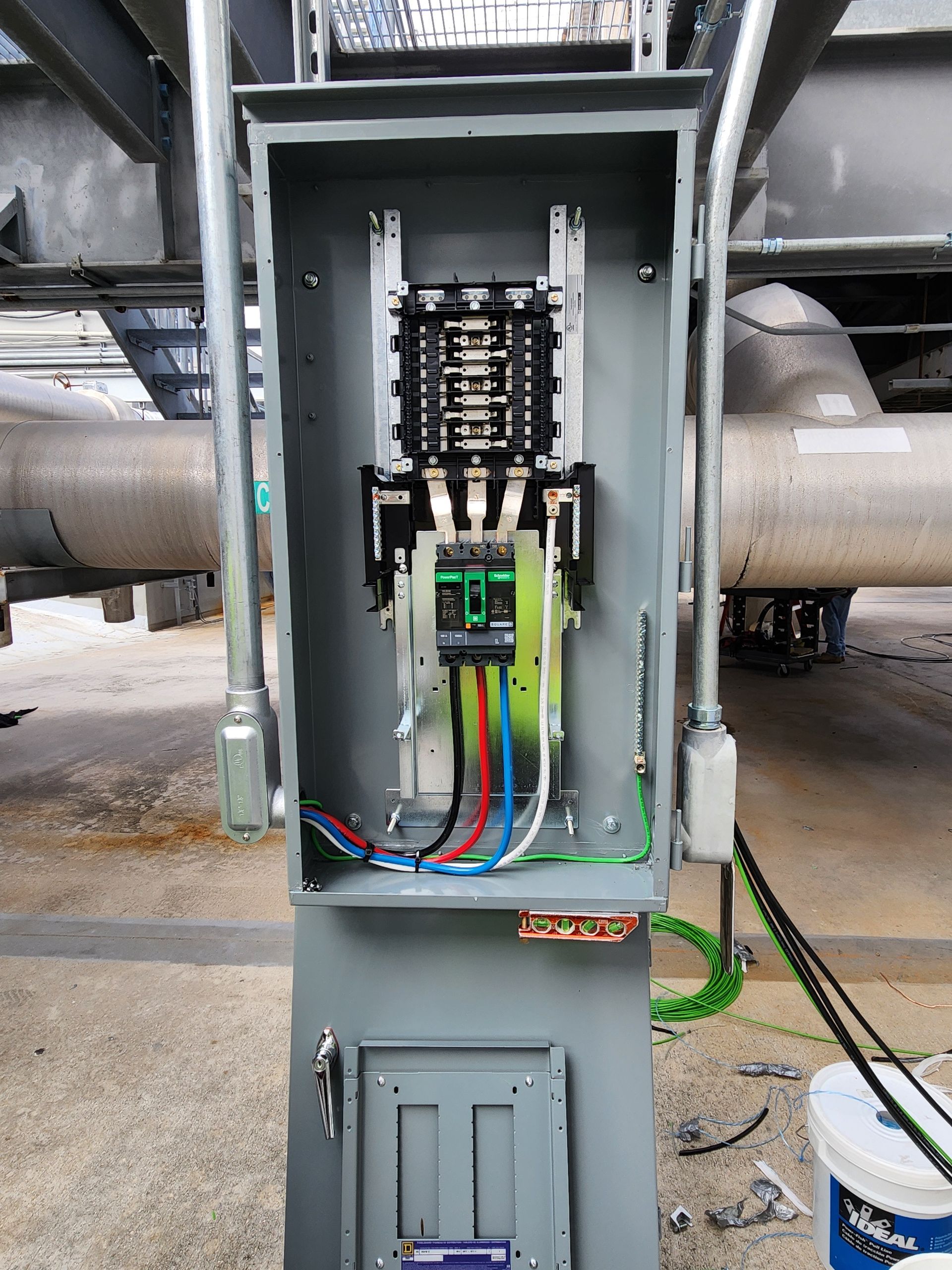 Open electrical panel with wires and circuit breakers, outdoors near pipes.