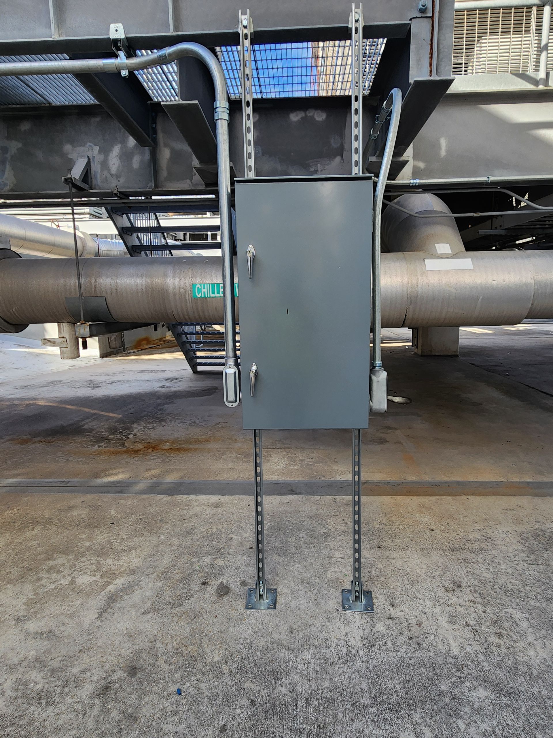 Gray electrical box on metal legs, mounted near pipes and industrial equipment.