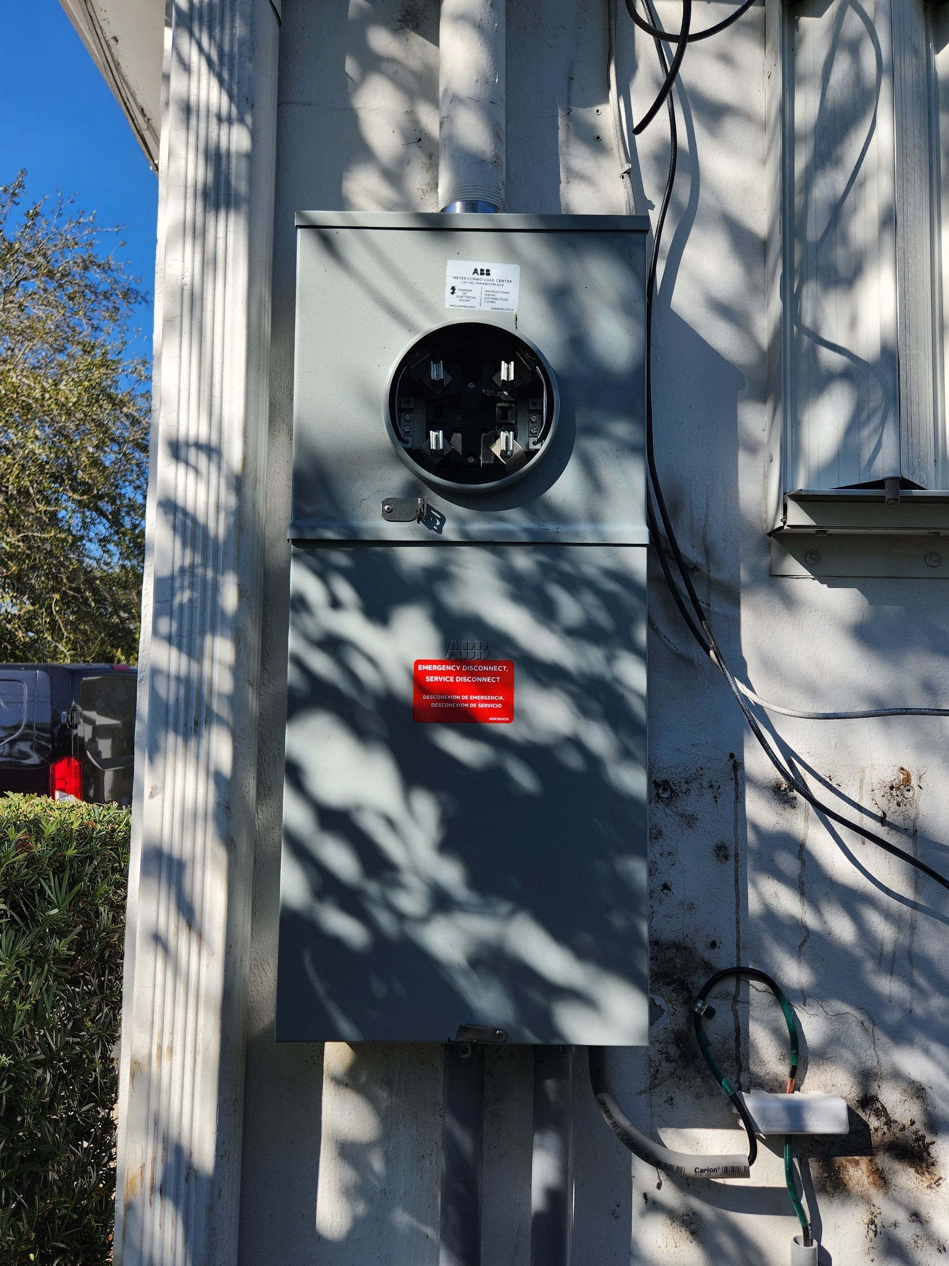 Electrical meter and panel box mounted on a white house exterior. Gray metal, black meter socket, wires.