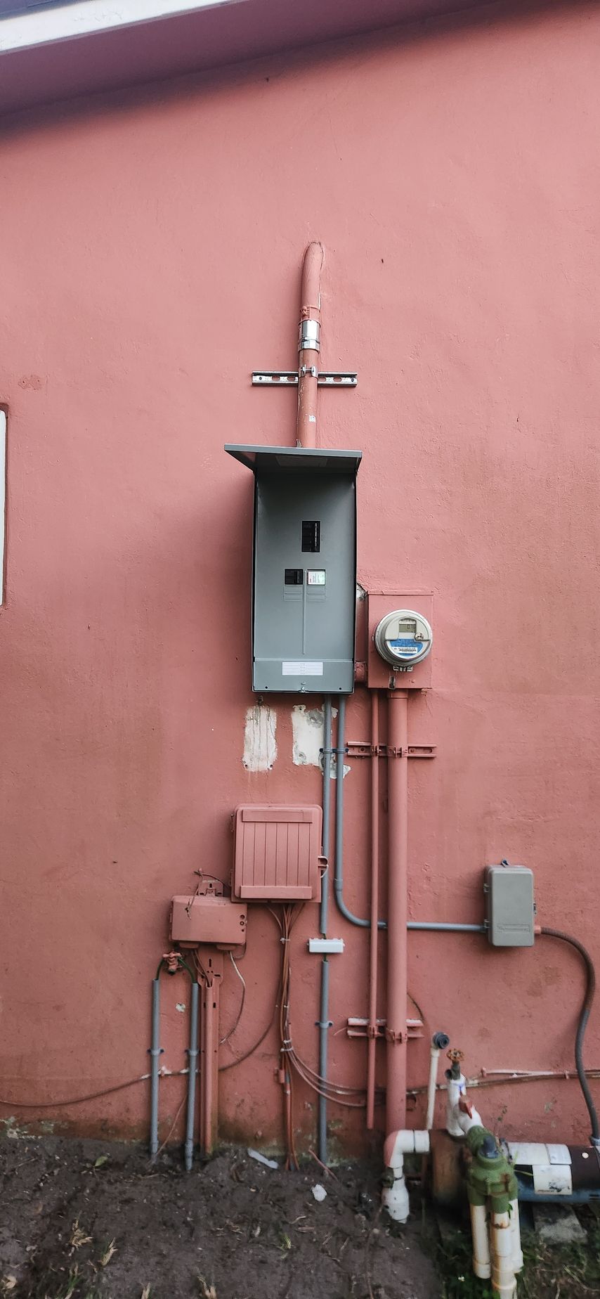 Electrical equipment on a pink building wall: electrical panel, meter, conduits, and junction boxes.