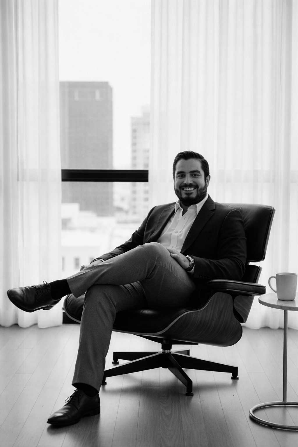 Hombre sentado en silla de oficina, con las piernas cruzadas, sonriendo, vestido con traje, entorno de oficina moderno.