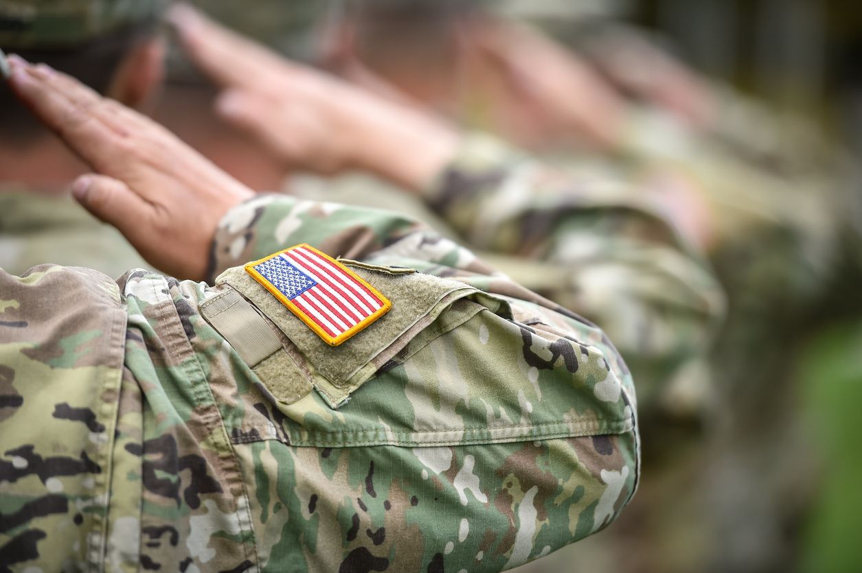 Soldiers in camouflage uniforms saluting, US flag patch on sleeve.