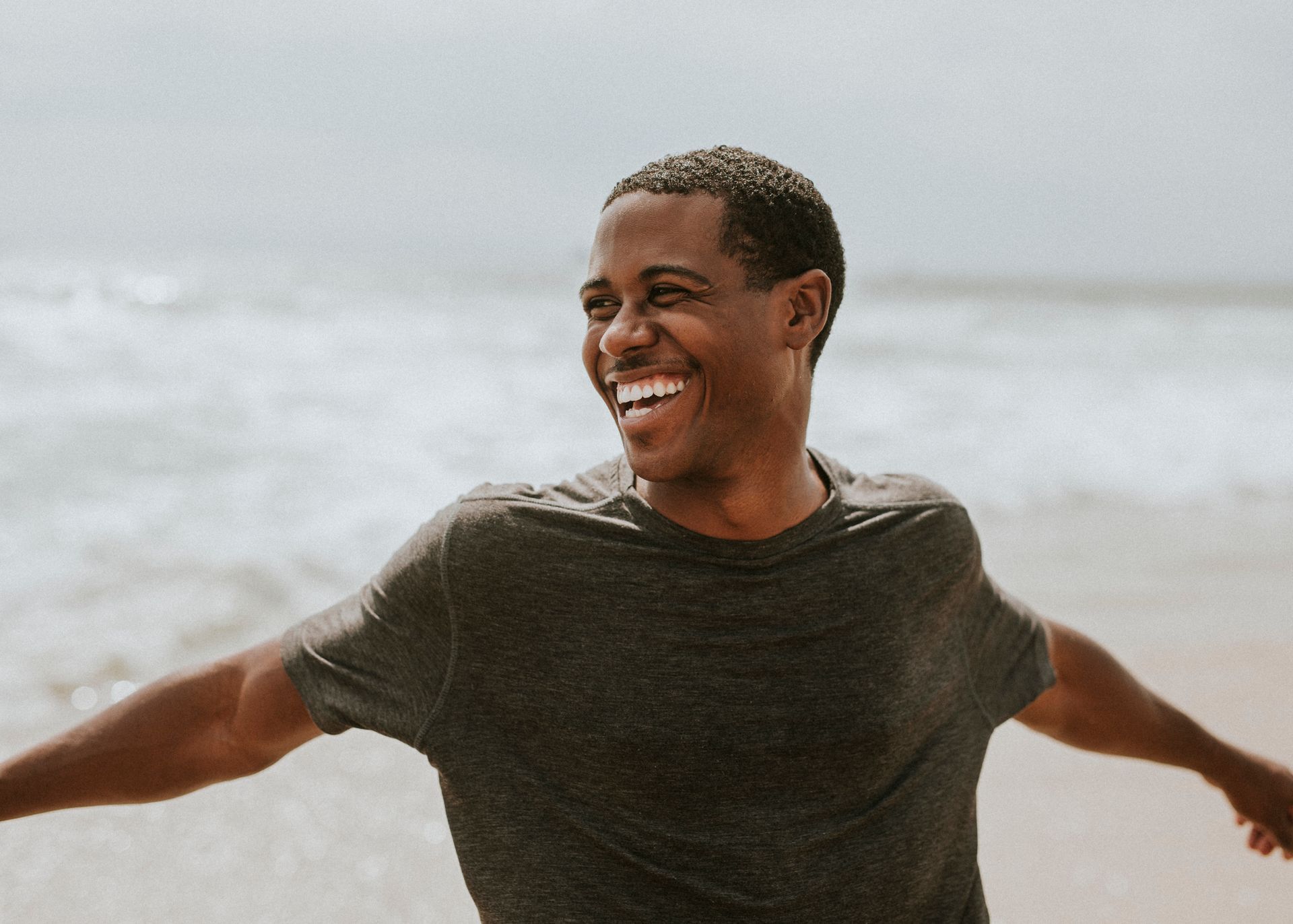 Man smiles with arms outstretched at the beach, waves in background.