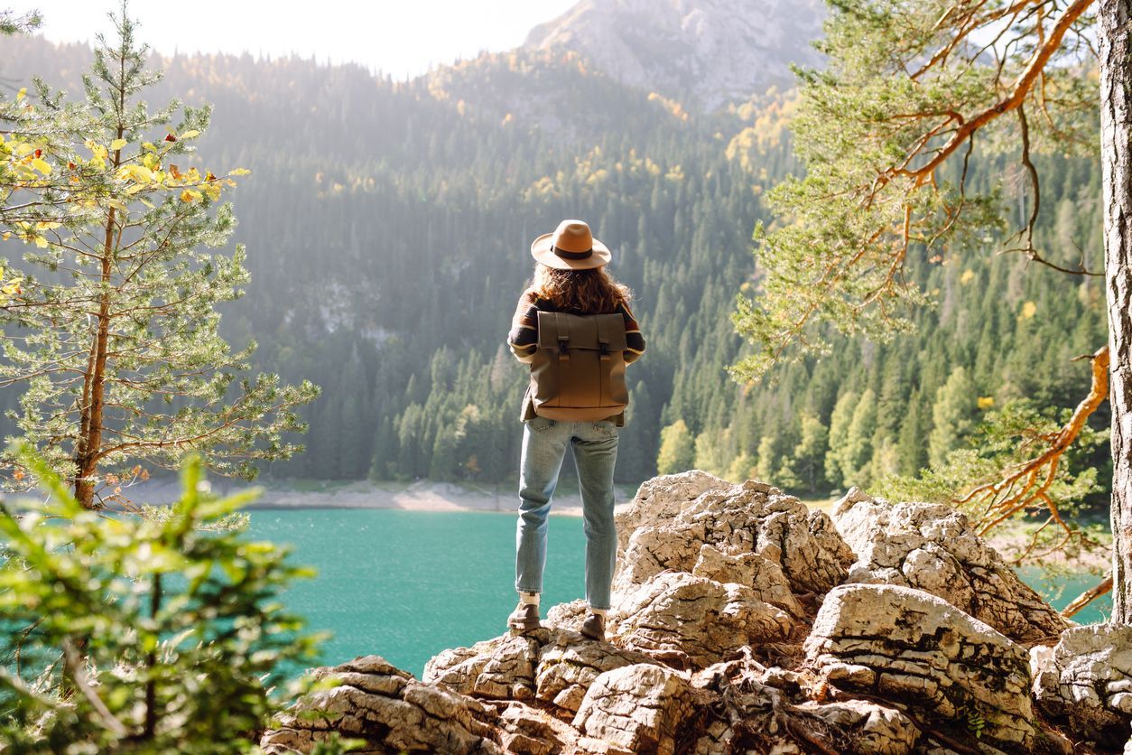 Woman with backpack stands on rocks, overlooking a lake and forest.
