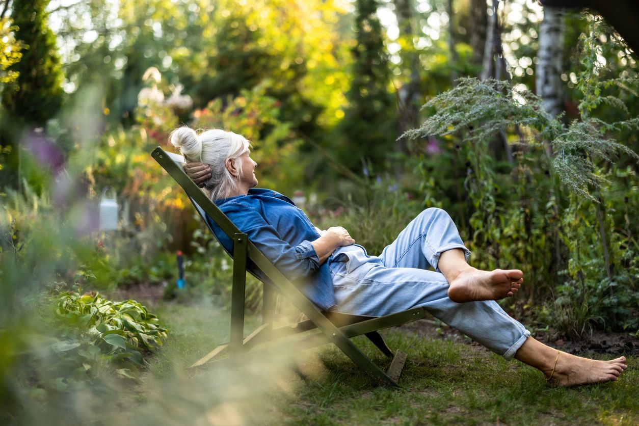 Woman relaxing in a folding chair in a sunny garden, barefoot, hands resting on abdomen.