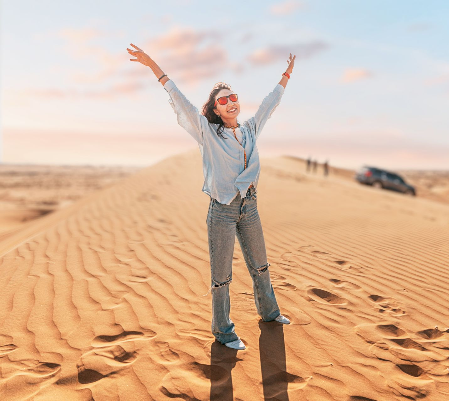 Woman stands on a desert dune with arms raised, smiling. A car and figures are in the distance.