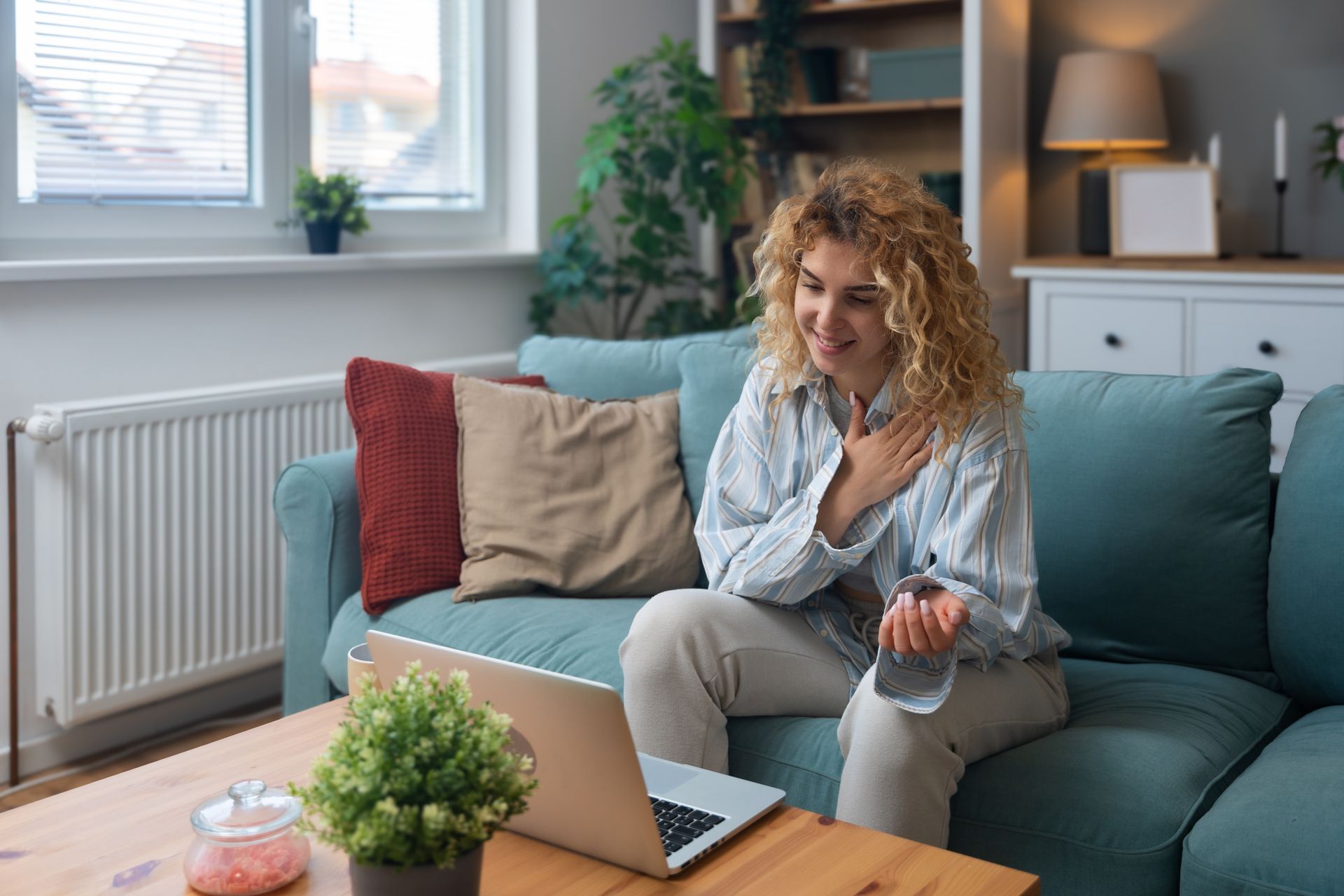Woman on couch smiling, using a laptop, gesturing, holding water glass, indoor setting.