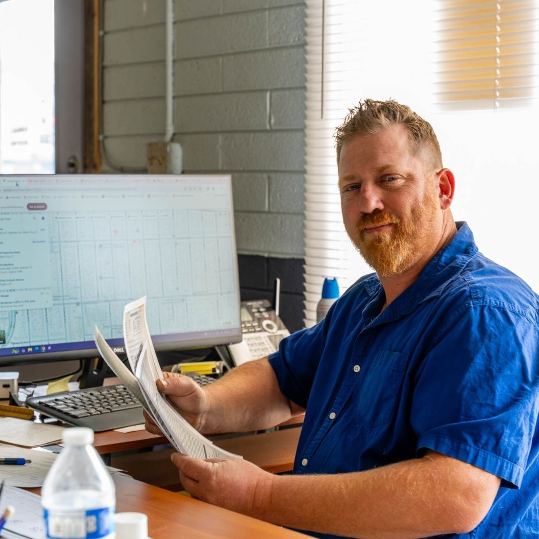Man in blue shirt at a desk holding papers, looking at the camera. A computer monitor is visible.