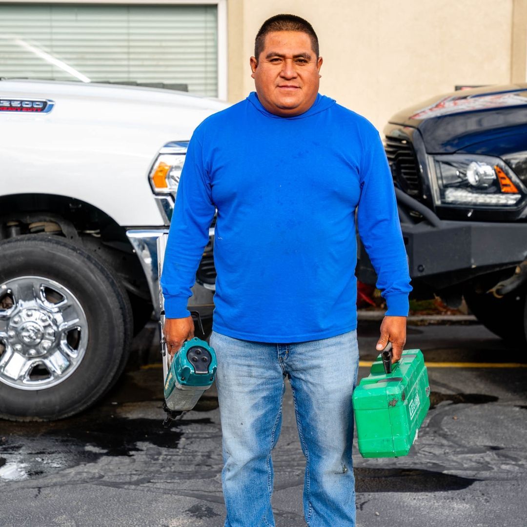 Man in blue shirt, holding tools, stands between two trucks.