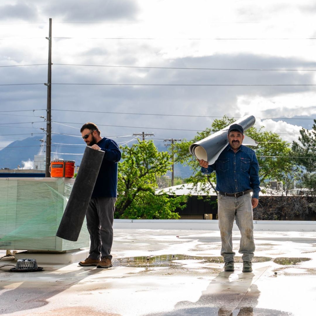 Two roofers on a flat roof, one holding a roll, the other a panel. Overcast sky with mountains.