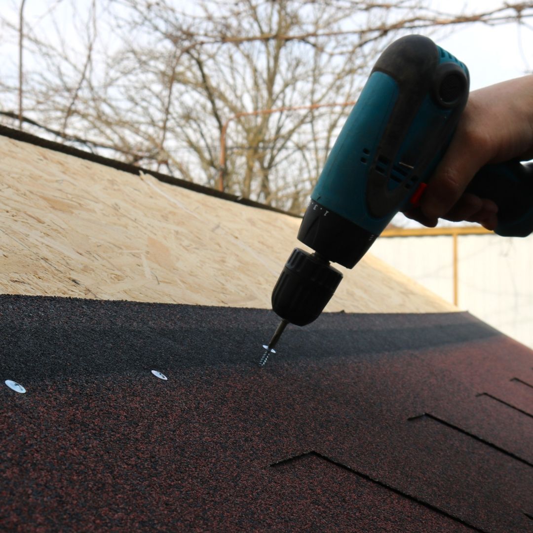 A person using a power drill to secure asphalt shingles on a roof.