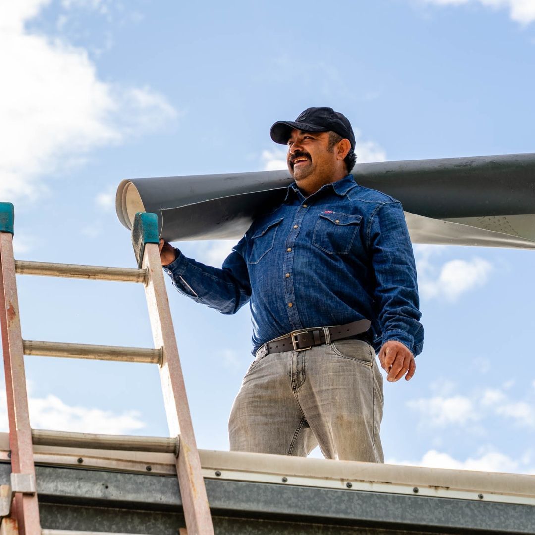 Man on ladder carrying rolled roofing material on a rooftop, sunny blue sky.