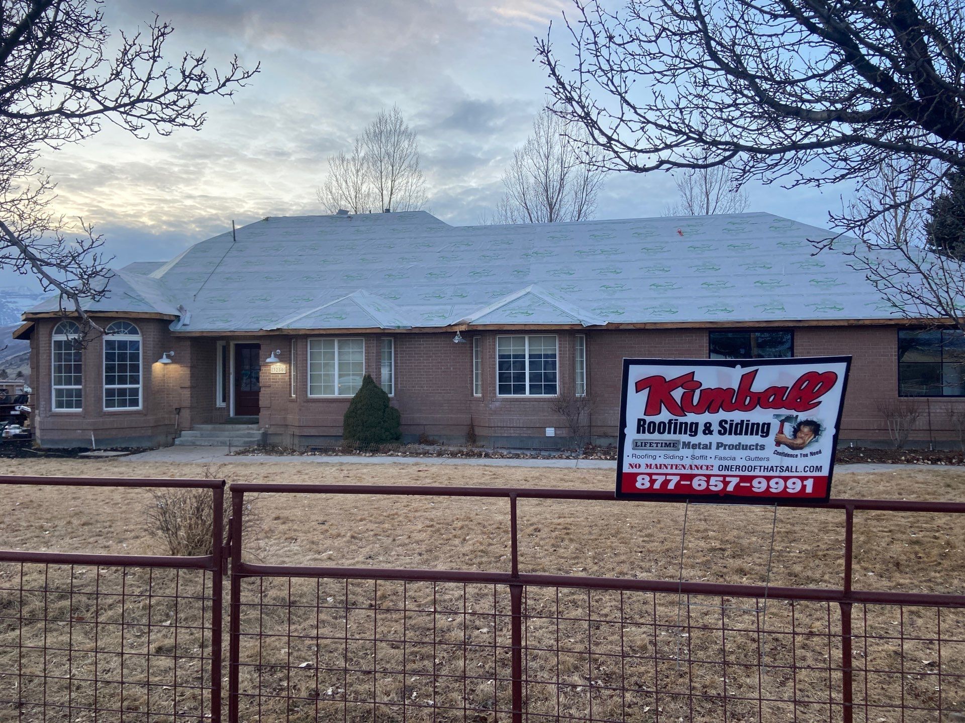 House with light gray roof, brown brick exterior, and a sign for Kimball Roofing & Siding in front of a brown fence.