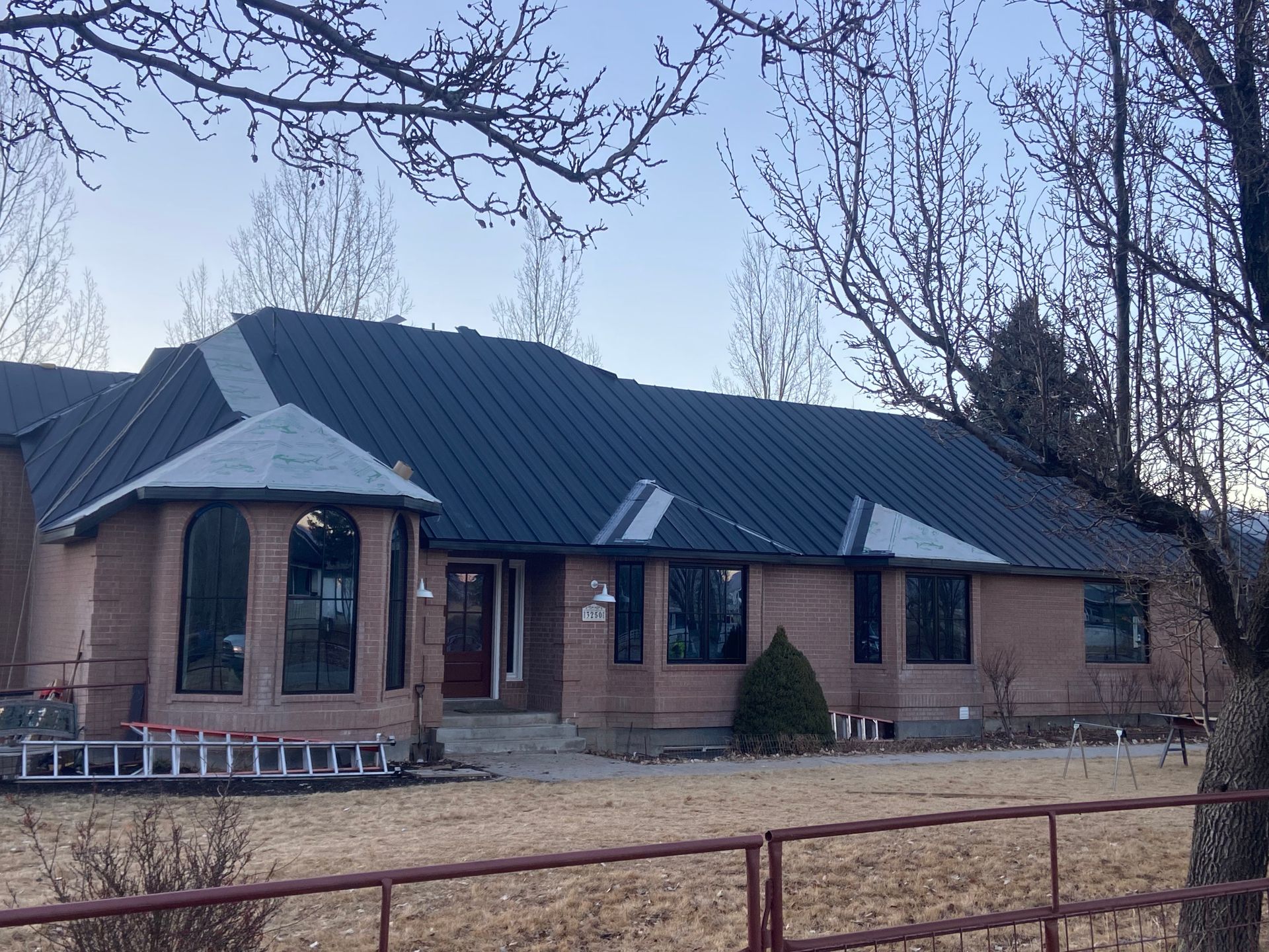 Brick house with dark metal roof, arched windows, and a ladder, behind a brown fence.