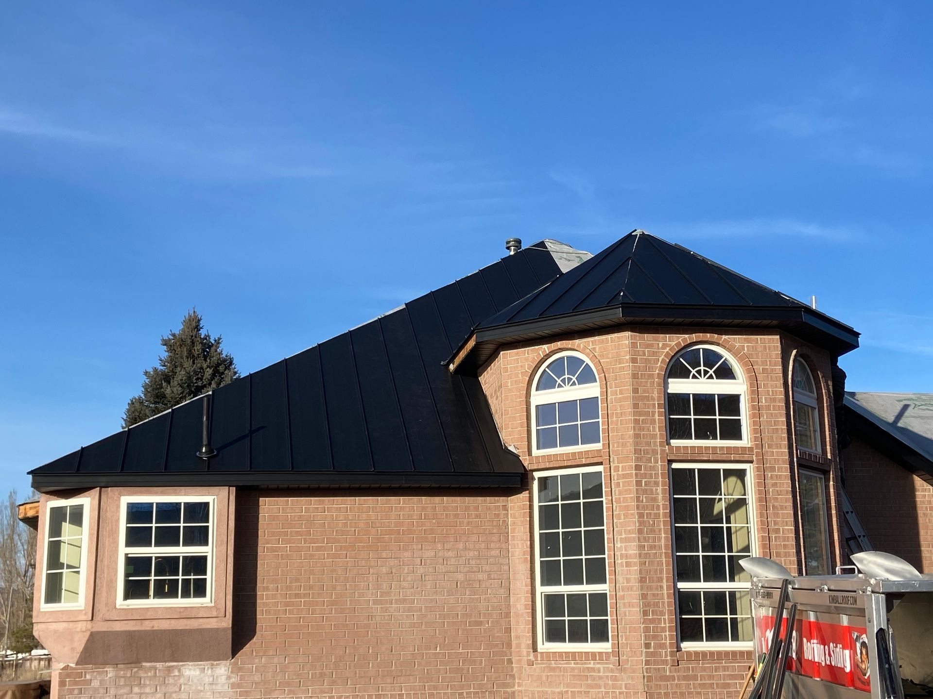 Black metal roof on a brick building with arched and rectangular windows under a blue sky.