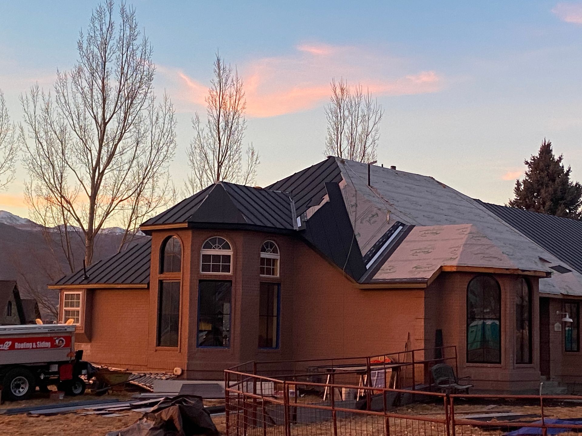 House under construction with dark metal roof, large windows, and unfinished stucco walls against a sunset sky.