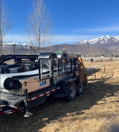 Trailer loaded with metal materials in a field, mountains in the background, under a blue sky.