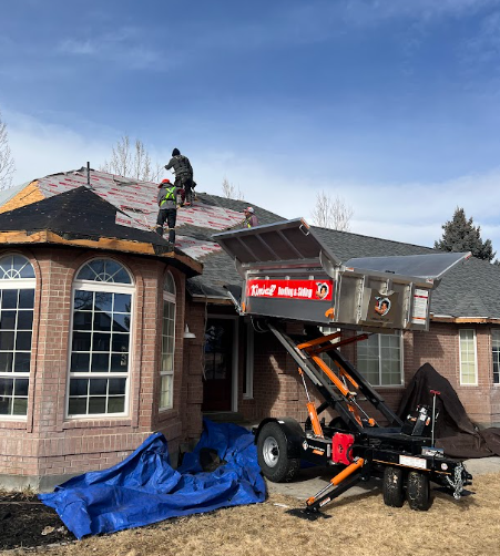 Roofers replacing shingles on a house with a mechanical lift. Blue tarp on the ground. Clear sky.
