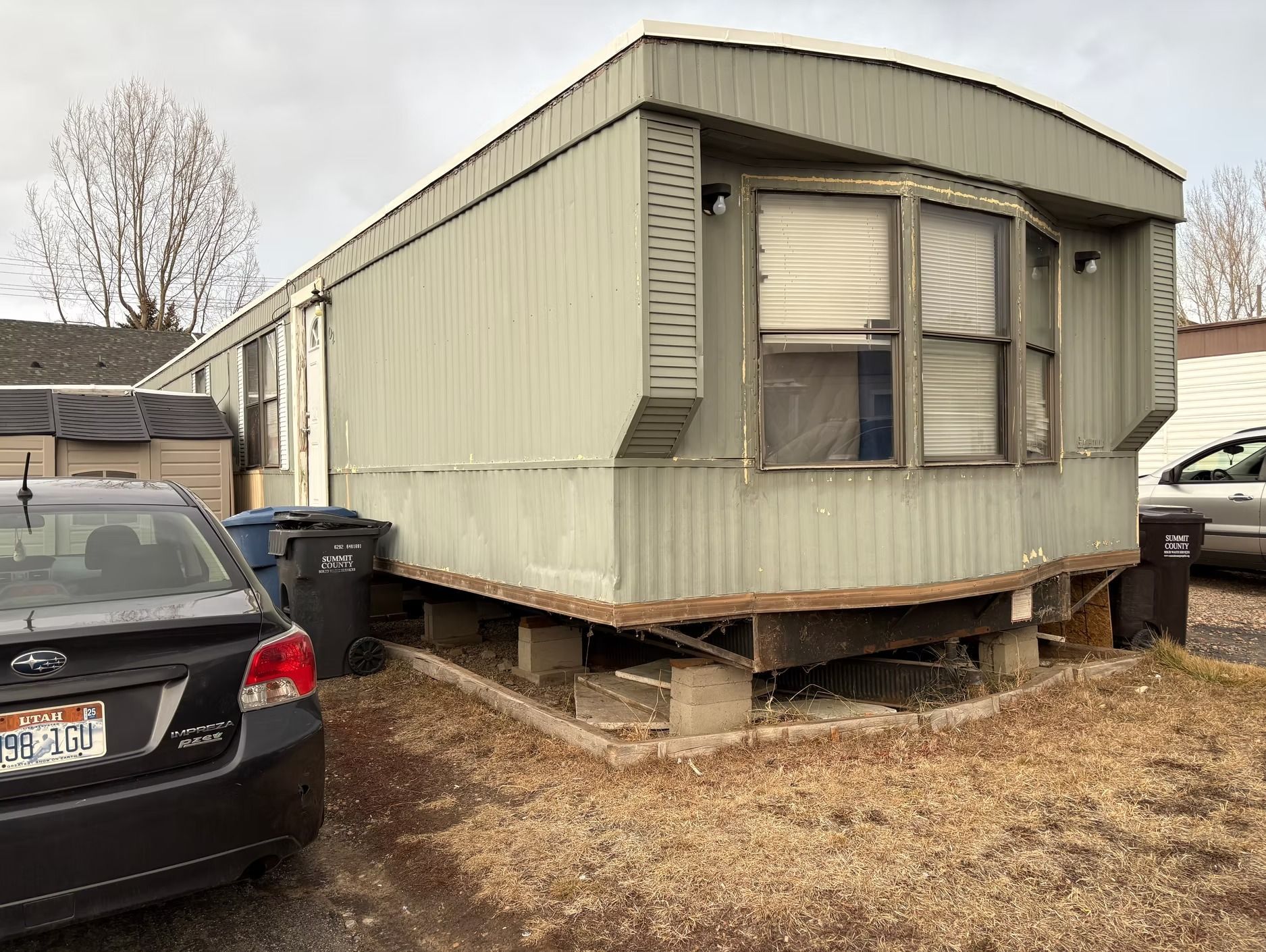 Mobile home, light green siding, with a car parked beside it on a cloudy day.