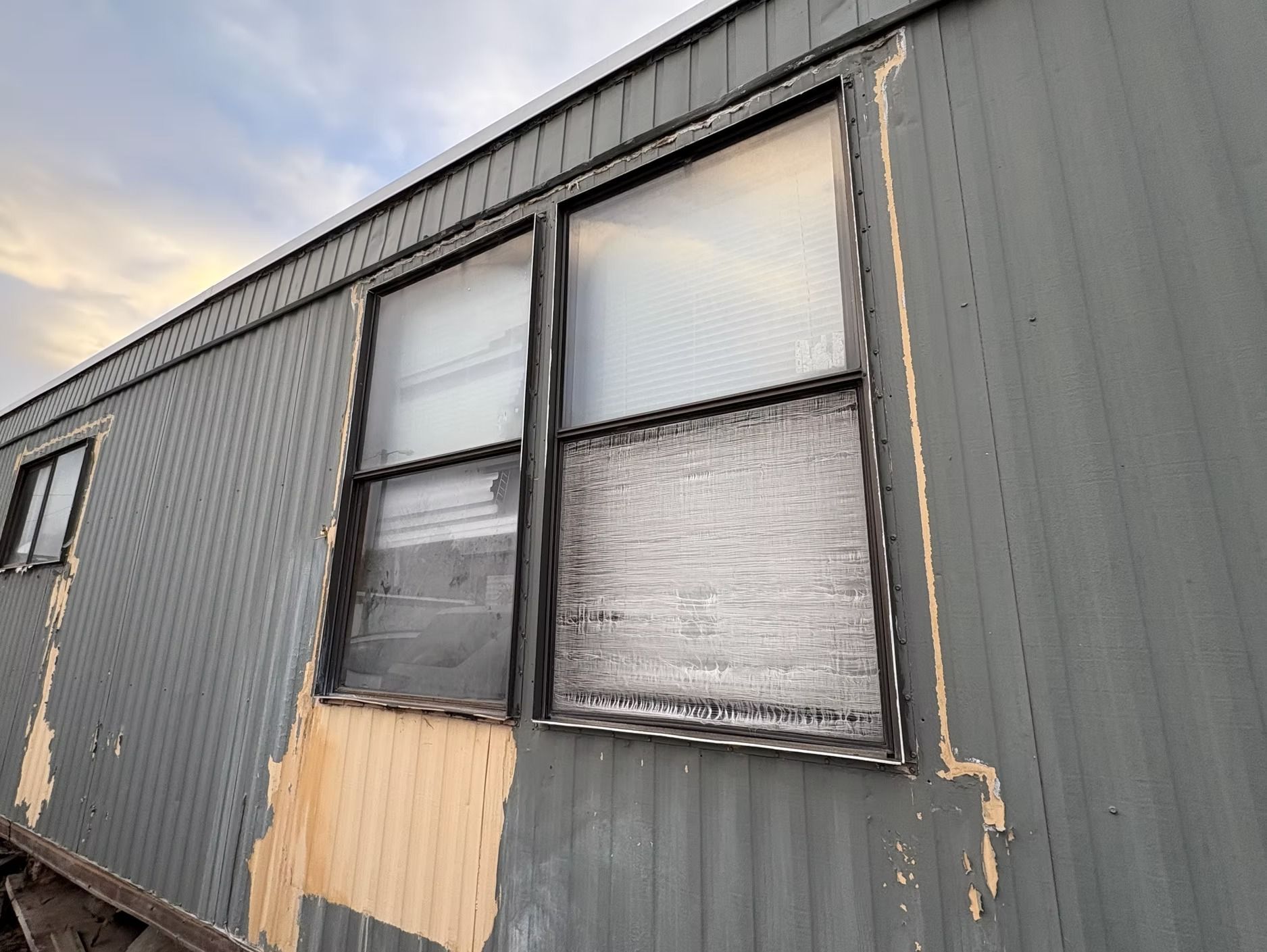 Weather-damaged mobile home exterior with peeling gray paint and frosted windows against a cloudy sky.