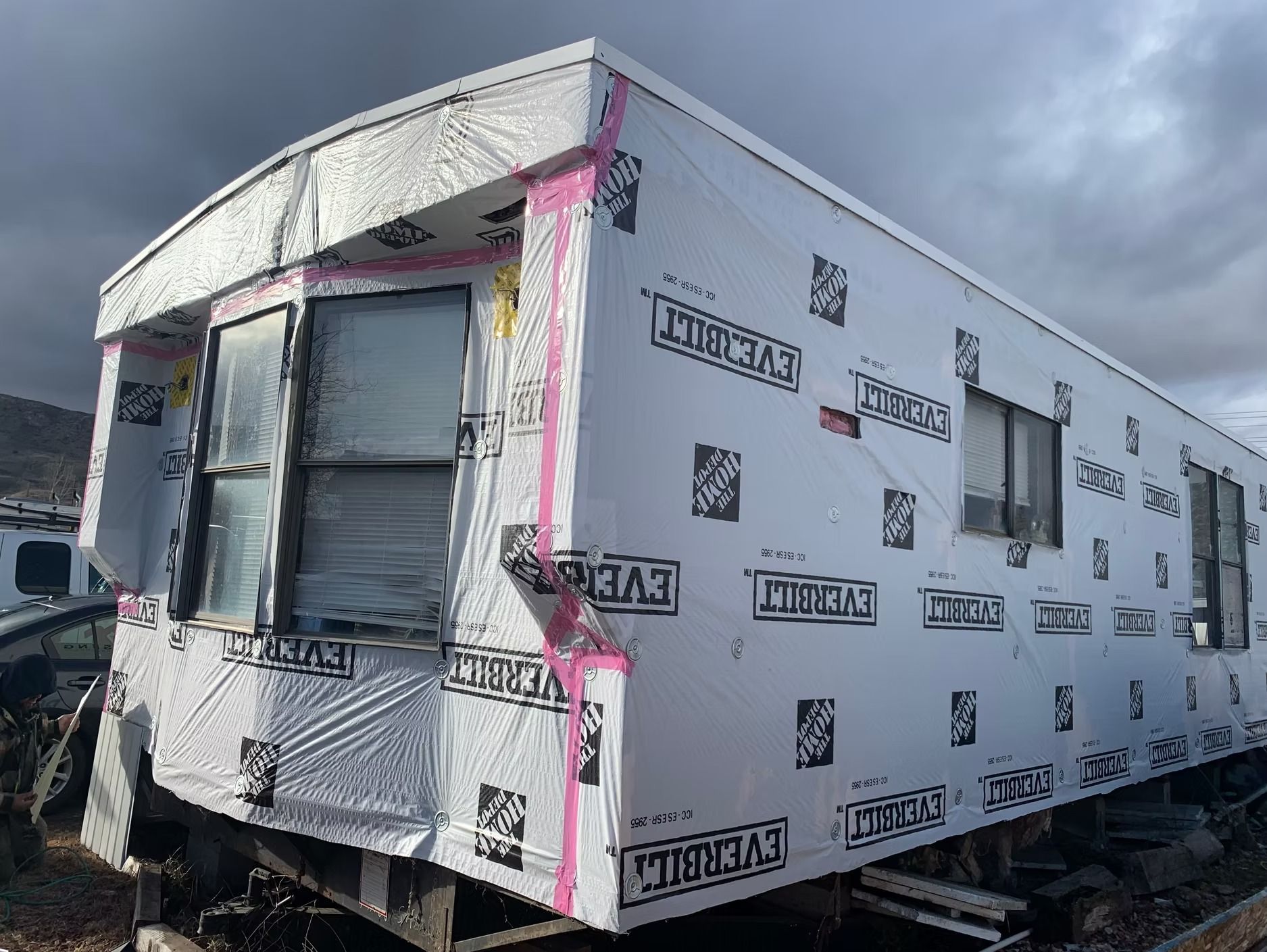 Mobile home exterior wrapped in white construction material; windows, pink tape, and overcast sky.