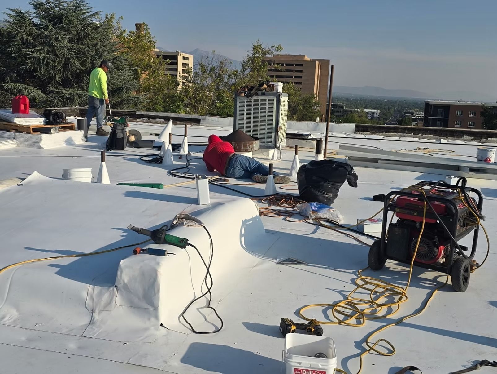 Workers on a flat roof repairing membrane, using tools, generator, and supplies. City and mountains in background.