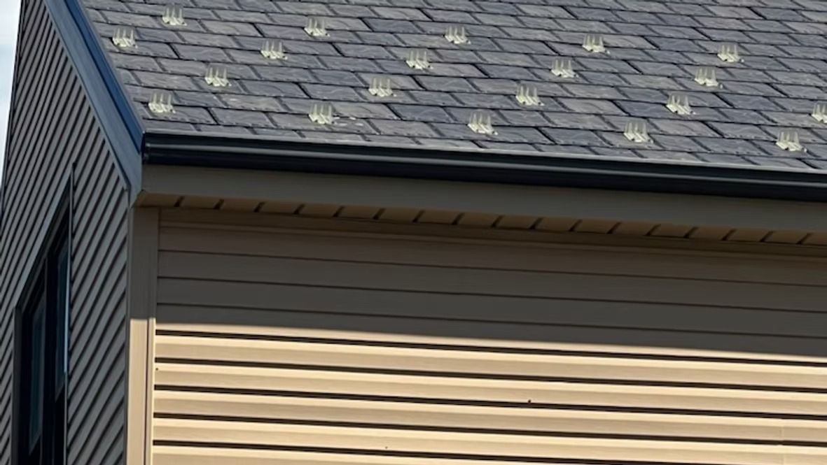 Corner of a building with brown siding and a dark gray shingle roof, black trim and gutter.