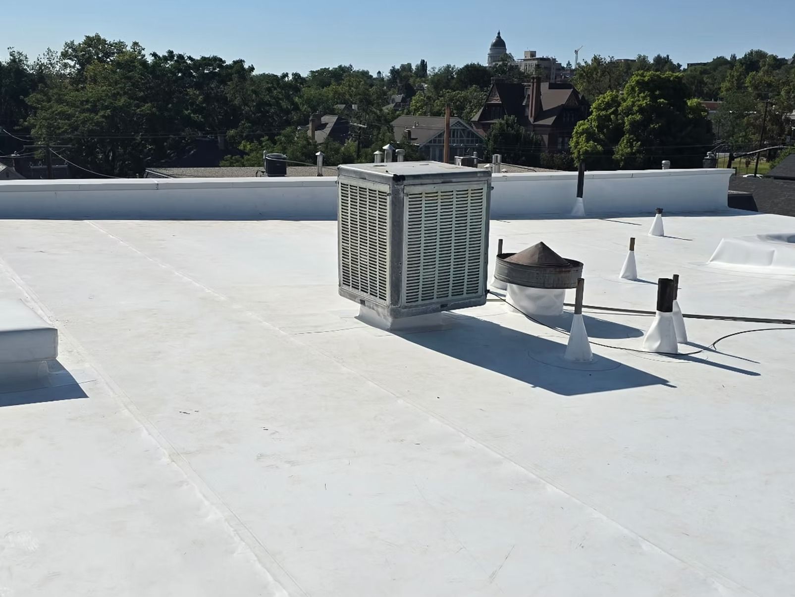 Flat white commercial roof with a large air conditioning unit. Buildings and trees in the background.