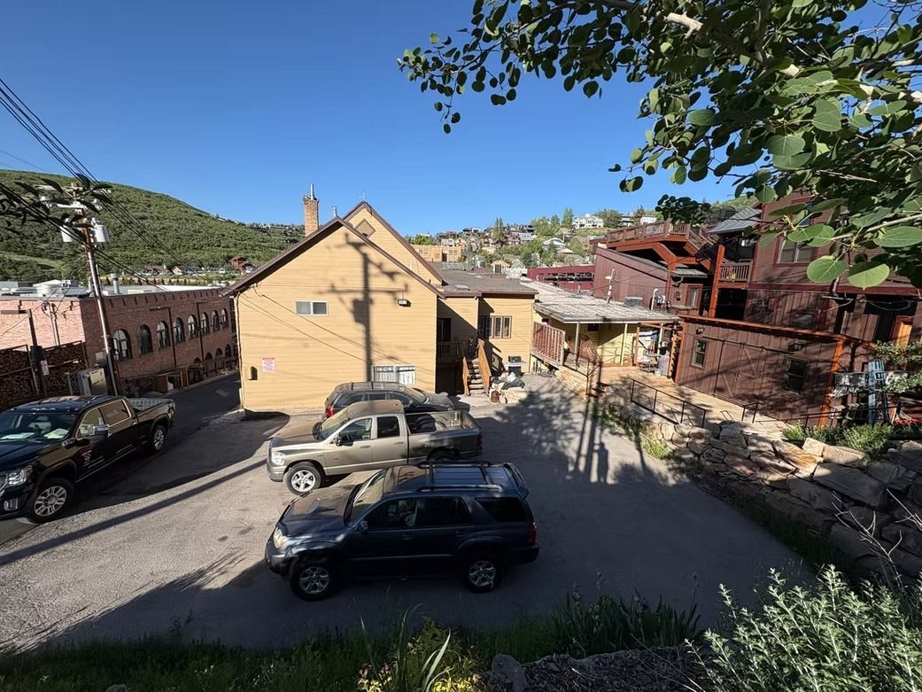 A street scene with buildings, vehicles, and mountain backdrop under a blue sky, trees in the foreground.