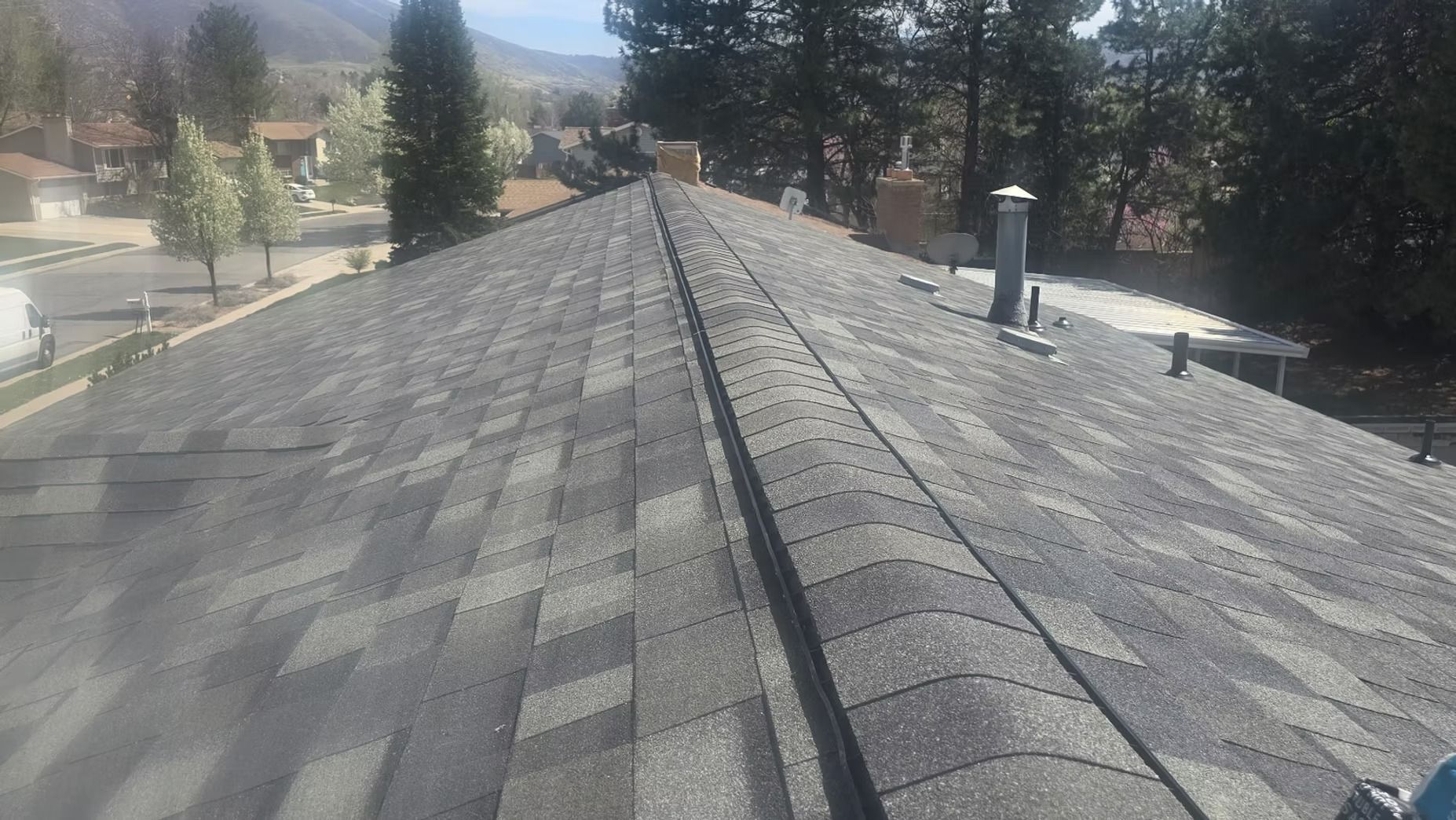 Gray asphalt shingle roof, with a ridge vent. Daytime, outdoor setting, residential neighborhood in the background.