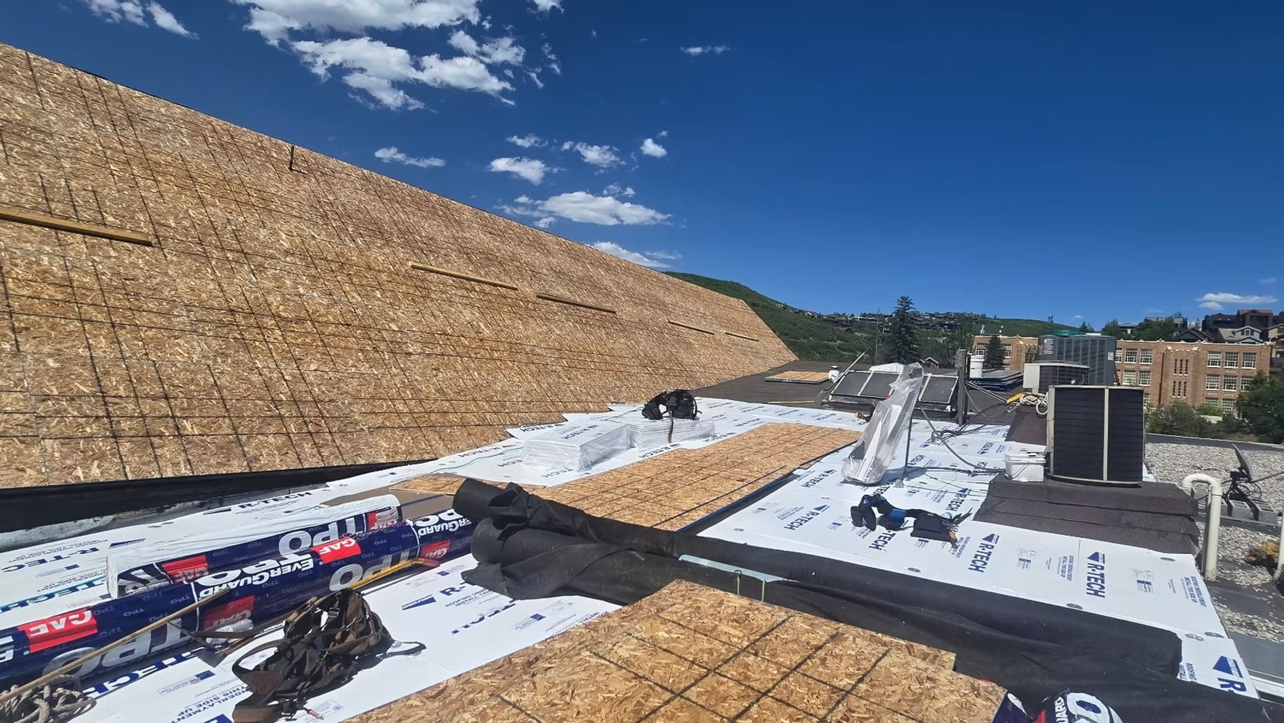 Rooftop under construction with plywood, black underlayment, and workers against a blue sky.